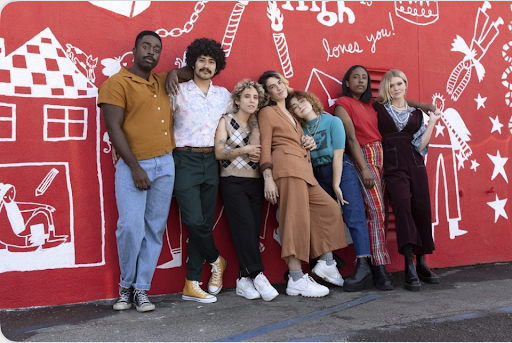 A group of seven people wearing colorful outfits stand outside in front of a red wall with white illustrations