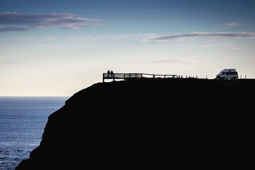 Couple on bench looking over sea with campervan parked nearby