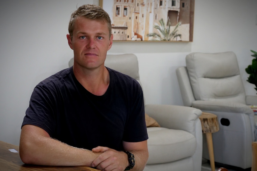 A blonde man sitting at a table with a sombre expression, he is wearing a black t shirt