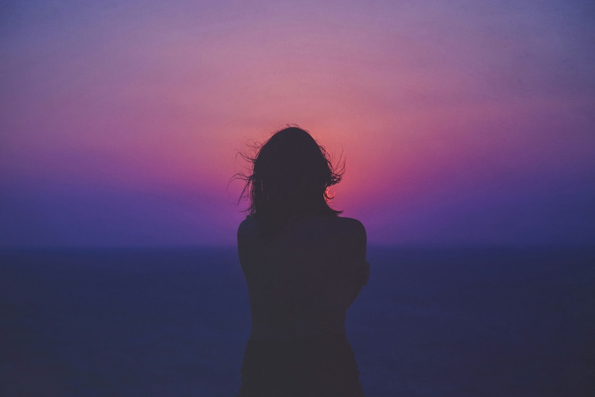 A woman is silouhetted from behind, her hair windblow, as she looks into a bright pink and purple sky.