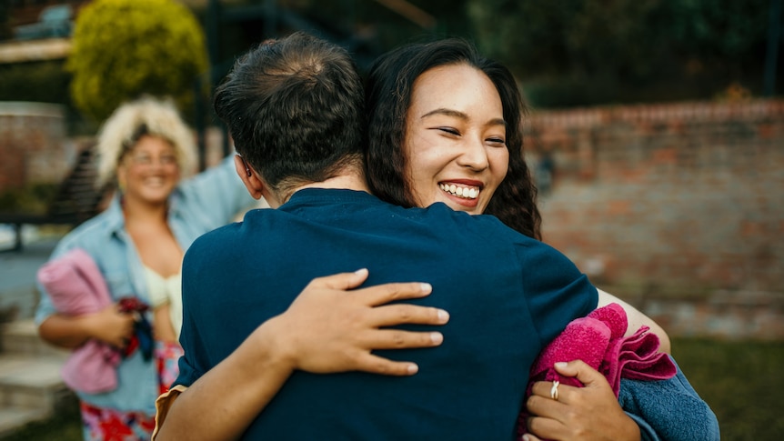 A young smiling woman embraces a young man, whose back is to the camera. Another smiling woman is visible in the background.