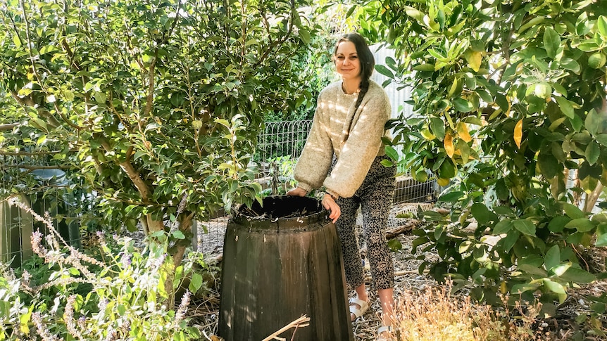 Koren Helbig smiles while standing next to a compost bin in a sunny, lush garden.