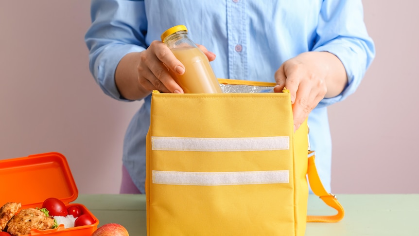 A woman puts a bottle of juice into an insulated yellow lunch bag ,a packed lunchbox sits beside it. Bringing lunch to work.