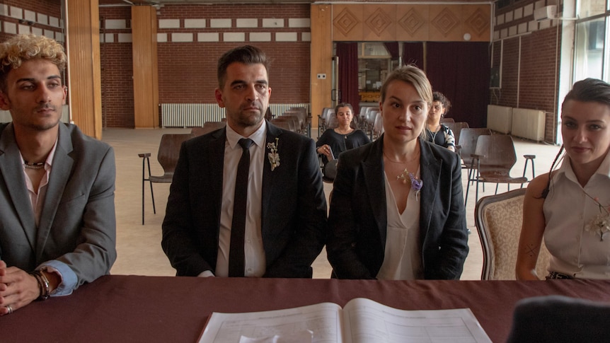 Four people - two men and two women - sit at a desk in suits and dresses, facing a panel of people.