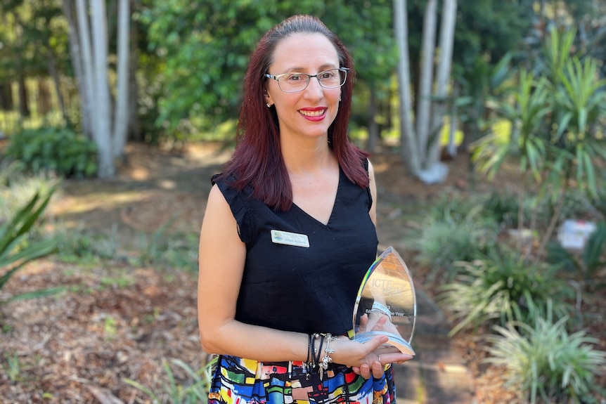 A female teacher with long brown hair stands in a garden area, holding an award and smiling.