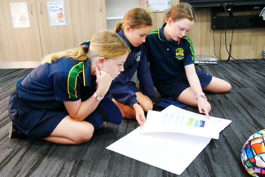 Three primary school girls sit on a classroom floor looking at ideas in a notebook.