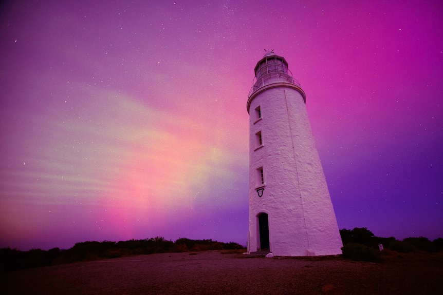 A lighthouse stands in front of a pink and purple sky lit by aurora