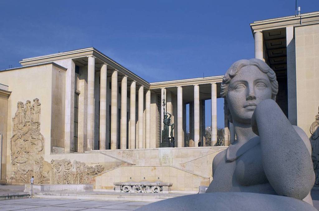 A columned building surrounding a plaza with a sculpture of a woman in front of it.