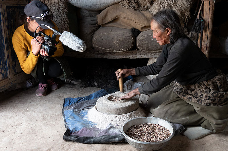 An older woman, seated on the floor, grinds flour in a stone mortar and pestle, while a younger woman kneels down, filming and recording her.