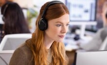 a person with red hair wears the  Jabra Evolve2 65 headset while looking at a computer screen. The setting it an office environment with other people working in the background at computer stations.