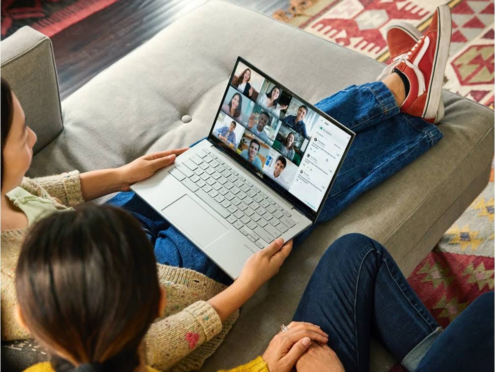 Two females sit on the same couch participate in a Google Meet meeting