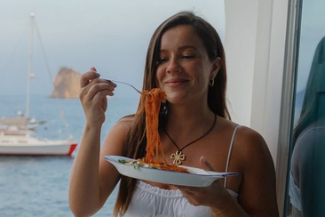 A girl sitting at a window with a sea view eating pasta.