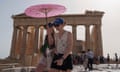 A young woman in a lace vest, shorts, baseball cap and sunglasses holds a cloth parasol over herself and the young woman next to her, also in shorts and a baseball cap, in front of the columns of the Acropolis in Athens during a heatwave on 12 June