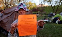 Weathered-looking white woman wearing pink and blue sunhat holds up bright orange piece of paper, with tents on green grass behind her.