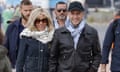 Emmanuel and Brigitte Macron walk side by side. The president is wearing a black baseball cap and a blue scarf tucked into a black bomber jacket. Brigitte is wearing sunglasses, a cream and black scarf and a denim jacket