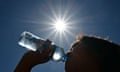 A figure in silhouette drinks from a clear blue-tinted water bottle, with a blazing sun above in a clear sky/