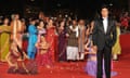 A man wearing a tuxedo stands on a red carpet. Behind him three dancers in saris kneel, smiling and making gestures with their arms, and behind them a group of people smile and throw rose petals.