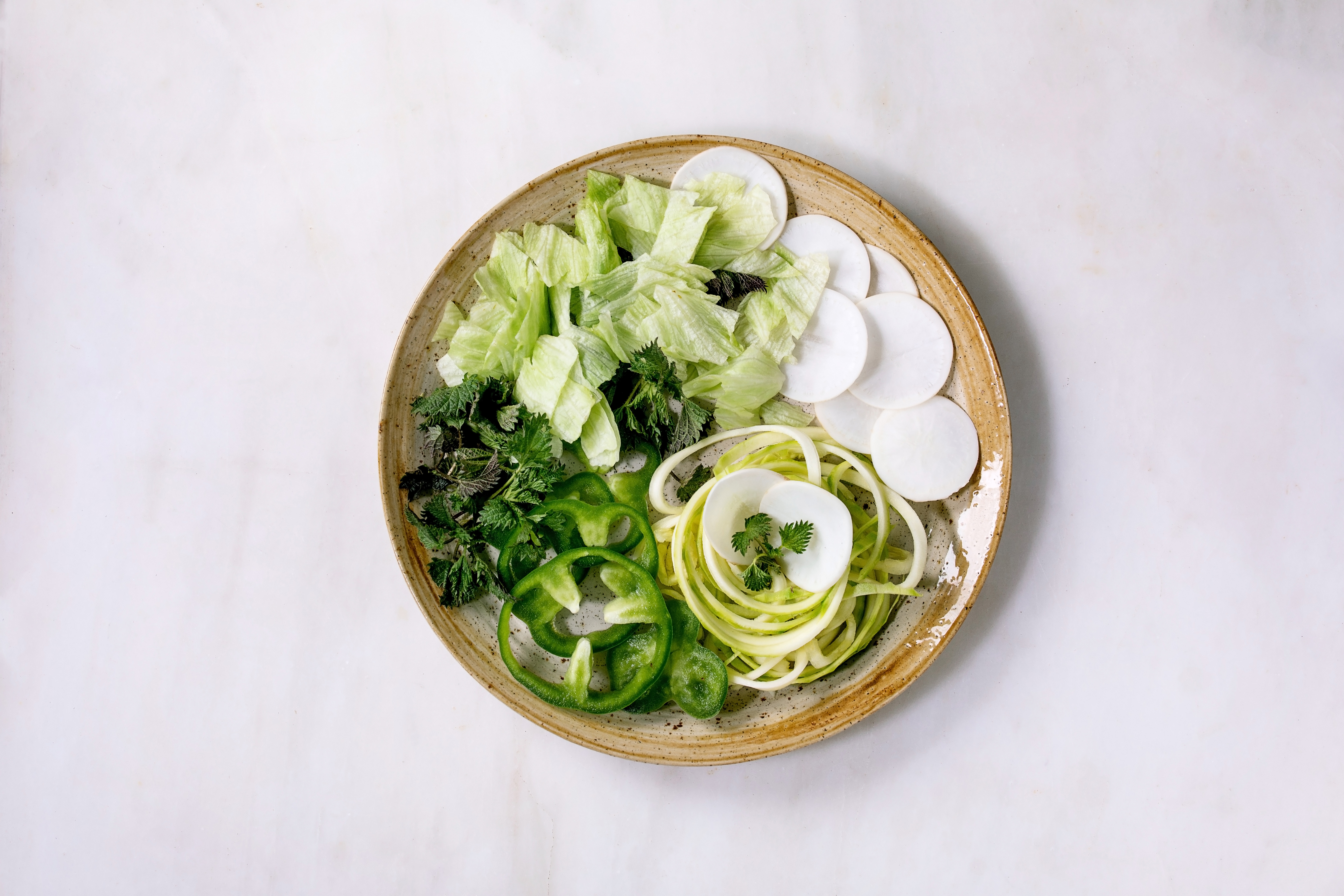 Fresh green raw vegetables and herbs spaghetti zucchini. white radish. green paprika. ice salad for cooking vegan dinner salad. Ceramic plate on white marble table. Flat lay. copy space