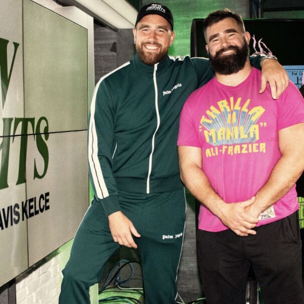 Travis and Jason Kelce standing beside 'New Heights' podcast promotional display