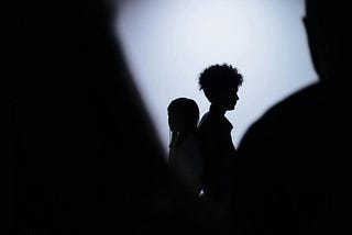 Photo of a silhouetted girl and boy standing back to back. In the foreground are silhouettes of two larger figures that seem to loom over the children — the one on the right appears to be an adult man.