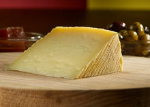 A wedge of cheese with a textured rind is placed on a wooden board, with a bowl of olives and red condiments in the background.