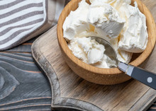 A wooden bowl filled with scoops of Mascarpone Cheese, placed on a wooden cutting board. A metal spoon with a black handle rests inside the bowl. A striped cloth napkin is partially visible in the background.