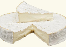 A wheel of Brie cheese, partially sliced, sits on a white surface with triangular piece of brie sits on the top of the wheel