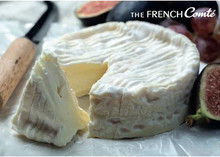 A disc of Camembert cheese, partially cut, with a triangular piece alongside, placed on a white surface with a knife and figs