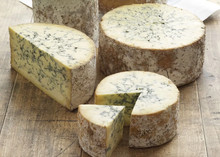 Wheels and wedges of stilton blue cheese on a wooden surface. The cheese wheels have a rough, natural rind, and the interior is creamy yellow with characteristic blue veins of mold running through it.