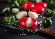 Small rounds of Babybel cheese with red wax, arranged on black marble alongside fresh parsley, green olives, and a cutting knife