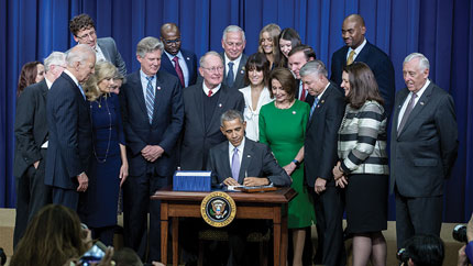 President Obama signing the Cures Act surrounded by Vice President Joe Biden and other legislators and senior White House officials.