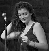 A black-and-white photo of Lorde performing. She is seen touching her hair and smiling while looking downwards