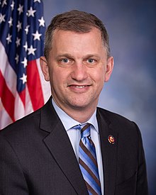 3/4 view portrait of Casten in a blue collared shirt. Parts of signs, others' heads, and background plant life are visible behind him.