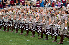 Row of students in uniform with knee-high boots. Each holds a bugle that dangles a pennant.