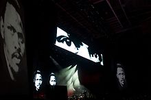 A darkened stage at a music gig. A small figure, barely visible, stands on the stage. The face of Biko, a young black man, is projected in black and white on some of the walls.