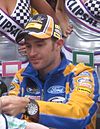 A man aged 30 at an autograph session. He is wearing a black, white and orange baseball cap, a blue and orange fleece and a watch on his left wrist