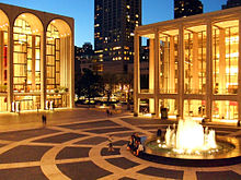 The corner of a lit up plaza with a fountain in the center and the ends of two brightly lit buildings with tall arches on the square