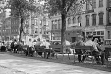 A dozen men in hats sitting on public benches facing an avenue of older stone buildings