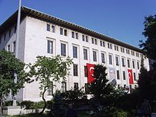 A four-story, white flat-roofed building with two Turkish flags and a portrait on the exterior