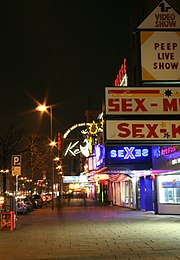 The Reeperbahn nightclub as seen from the sidewalk with its entrance lighted. Theatre marques say "Sex" and "Peep live shows".