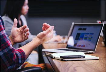 A meeting happens at a table, a paper and phone sits in the foreground, an open laptop in the background