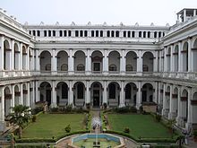 A white two storied building with arches and a courtyard in the foreground