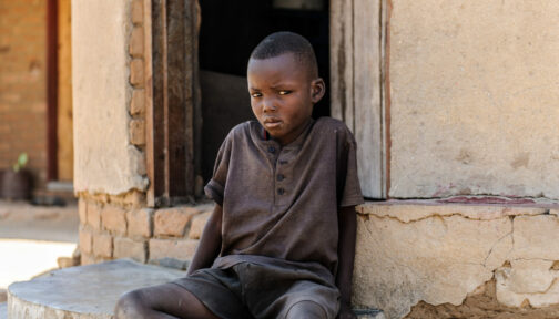 A young boy, Bretty, looks at the camera while sat on the ground outside a doorway.