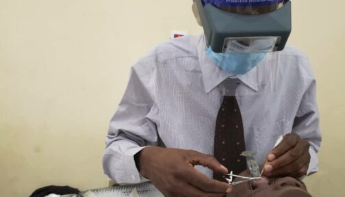 A man wearing a face mask, shield and optical loupes operates on the eye of a dummy