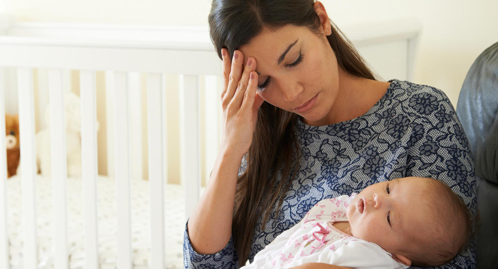 New mum sitting next to a baby cot, holding her head in pain while she looks at her sleeping baby in her lap