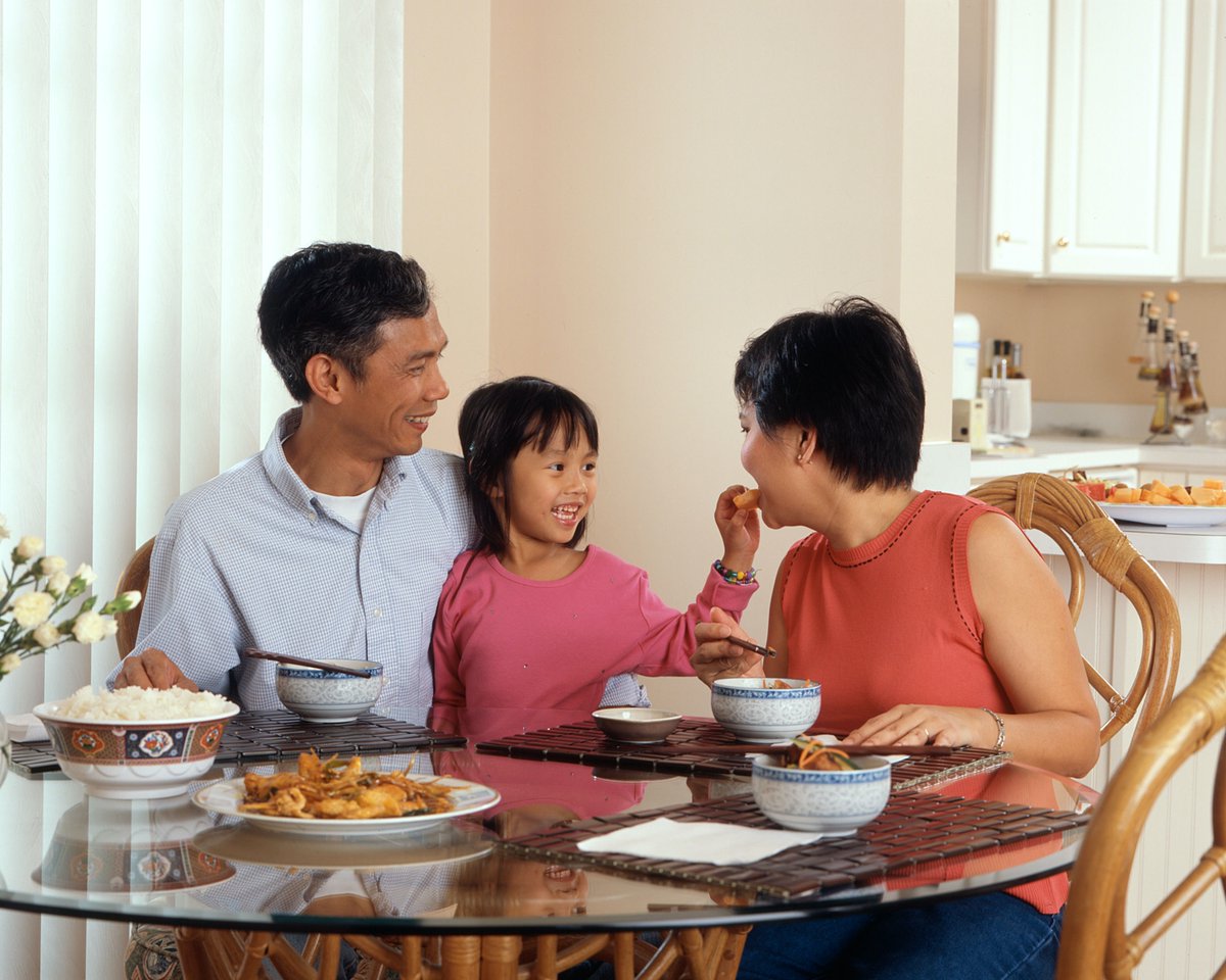 family sitting at table eating meal, child is putting a piece of food in mother's mouth