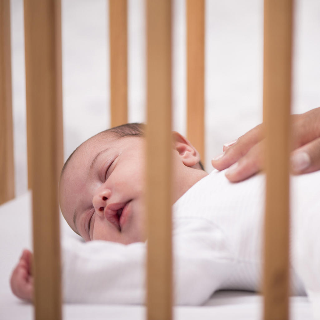 newborn sleeping on the back in his crib while the hand of grown woman is comforting him