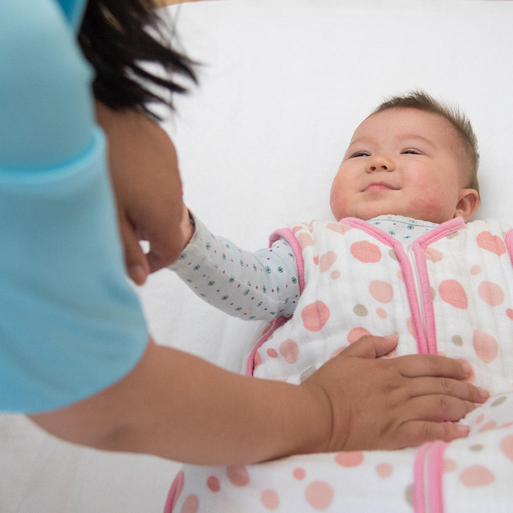 baby wearing a sleep sack while laying on her back in a crib being comforted by the hand of her mother