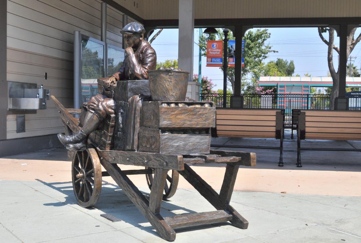 Photo of a bronze sculpture of a boy in a newsboy cap sitting atop a wooden cart with various pieces of luggage, fruit, etc on it. Photograph by Bryan Costales.