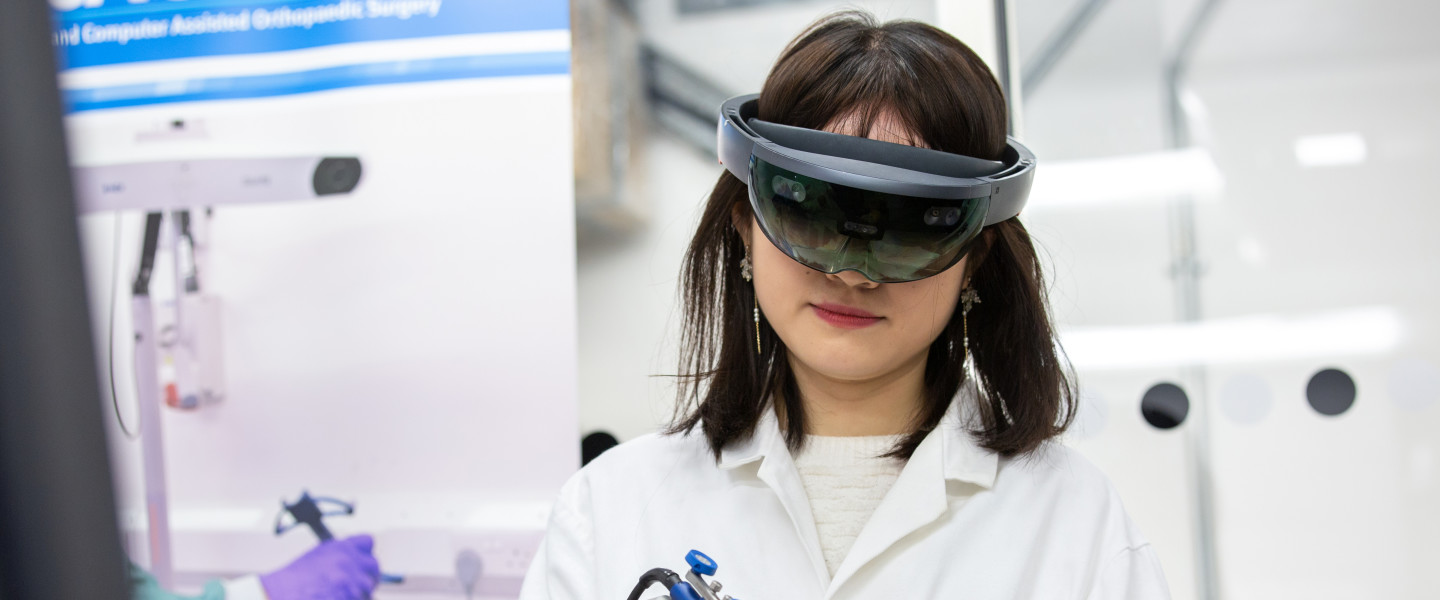 Light-skinned woman wearing black goggles uses a power tool in a lab. 